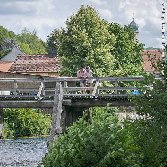 An der Balustrade der Holzbrücke vor Essing lehnt sich ein Pärchen an und schaut flussaufwärts. Hinter  ihnen sieht man einige Hausdächervon Essing umrahmt von Bäumen und deren Blätterdach.