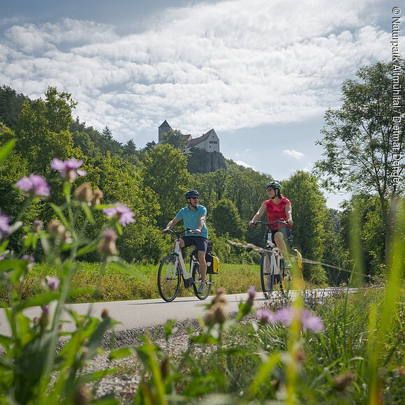 Zwei Radler, eine Frau mit weißem Helm und roten Shirt und ein Mann, neben ihr, ebenfalls mit Helm und blauen Shirt sind auf dem Radweg unterwegs. Die Zwei radeln und hinter ihnen ragt die Burg Prunn, die umgeben von Wäldern auf einem Felsen. Der Himmel scheint blau, nur ein paar Wolken verirren sich.