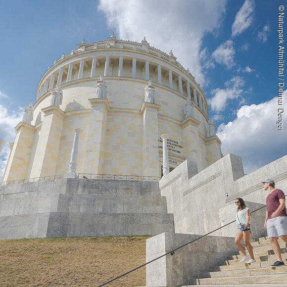 Zwei Frauen und ein Mann, in sommerlicher Kleidung, steigen die Treppen der Befreiungshalle hinunter und werfen einen letzten Blick zu ihr hinauf. Der Himmel strahlt blau und ein paar Wolken sind zu sehen.