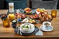 Der braune Tisch ist mit Kerzen, Orangensaft, Latte Macciatto, Weißwurst im Topf und Wurst, Käse reichlich gedeckt.