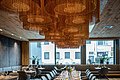 Eine lange Tafel  mit bequemen dunklen Stühlen ist aufgebaut. Im Hintergrund ist eine große Fensterfront. Von der Decke hängen braune stylische Lampen.