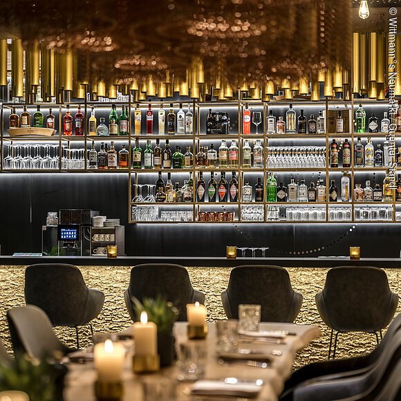 Wittmann°s Neumarkt Die Bar nimmt den größten Bereich ein mit dem offenen, gut gefülltem Regal. Davor stehen dunkle bequeme Bistrostühle. Ein Tisch mit Kerzen ist noch zum Teil zu sehen.