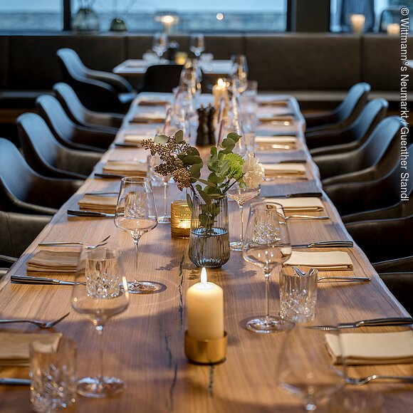 Restaurant im Wittmann°s Neumarkt Eine lange Tafel mit Weingläser, Besteck und brennenden Kerzen ist im Mittelpunkt. Die gemütlichen schwarzen Stühlen. Im Hintergrund ist eine große Fensterfront.