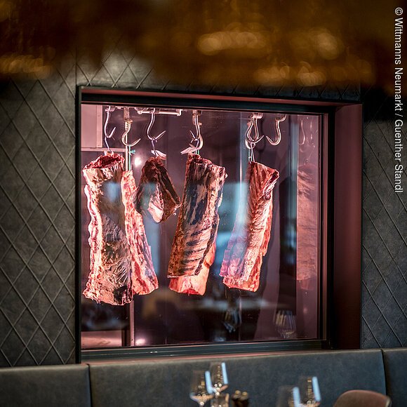 Restaurant im Wittmanns Neumarkt Im Restaurant ist ein Fenster in der Wand eingebaut. Durch dieses kann man einen Blick in den Kühlraum der Metzgerei mit Rinderhälften werfen. Diese reifen hier weiter. Ein Tisch steht genau vor dem Fenster und eine Sitzbank mit grauem Stoff ist an der Wand befestigt.