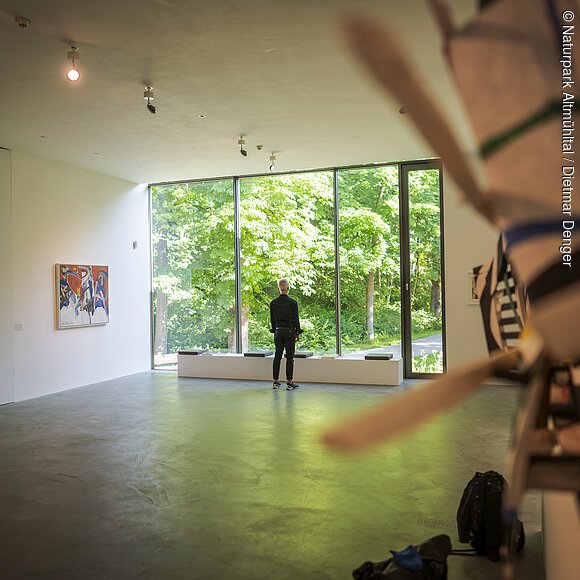 Ein Mann steht vor einer großen hellen Glaswand. Rechts und Links sind Exponate an der Wand zu erkennen.