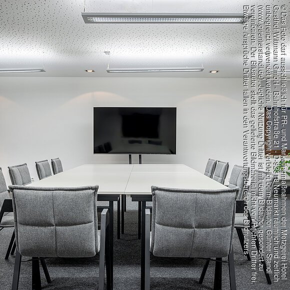 Der kleine Konferenzraum im Wittmanns Neumarkt Konferenzraum mit weißem Tisch, zwölf grauen Stühlen, großem Bildschirm und Pflanze am Fenster.