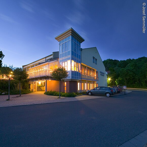 Beleuchtetes modernes Gebäude mit Parkplätzen und Bäumen bei Abenddämmerung unter klarem Himmel.