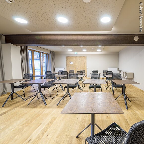 Leerer Schulungsraum mit Holzfußboden, mehreren Tischen und schwarzen Stühlen, Deckenleuchten und Vorhängen.