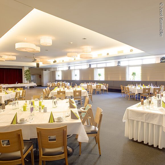 Großer heller Veranstaltungsraum mit eingedeckten Tischen und Holzstühlen, vorbereitet für ein Bankett.