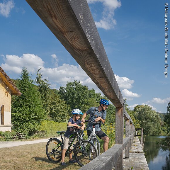 Familie mit vier Personen und Fahrrädern neben einem alten Haus und einem Fluss unter blauem Himmel.